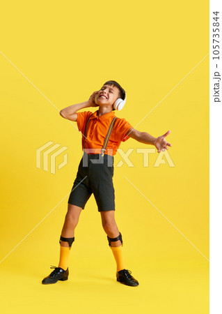 Full-length of little boy, child in stylish clothes listening to music in headphones against yellow studio background 105735844