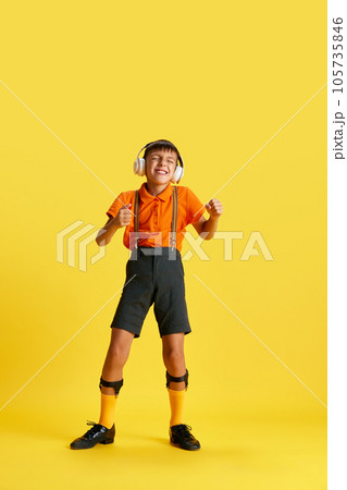 Full-length of little boy, child in stylish clothes listening to music in headphones against yellow studio background Full-length of little boy, child in stylish clothes listening to music in headphones against yellow studio background 105735846