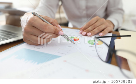 Female hand holds pen on graphs with indicators next to glasses. Female hand holds pen on graphs with indicators next to glasses. 105739305