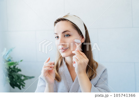 Young woman in bathrobe looking in mirror and making face massage with jade roller in the bathroom. The concept of natural procedures for skin care. Home Beauty self-care treatment. Selective focus Young woman in bathrobe looking in mirror and making face massage with jade roller in the bathroom. The concept of natural procedures for skin care. Home Beauty self-care treatment. Selective focus 105741366