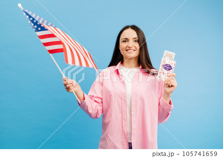 Young brunette woman in pink shirt poses on blue background in studio with american flag and hundred dollar cash with passport. Concept of success, travel, knowledge of foreign language 105741659