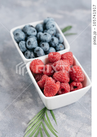 Top view on fresh pile of blueberries and raspberries in white square bowl on grey background. Healthy eating. Vegan food Top view on fresh pile of blueberries and raspberries in white square bowl on grey background. Healthy eating. Vegan food 105741917