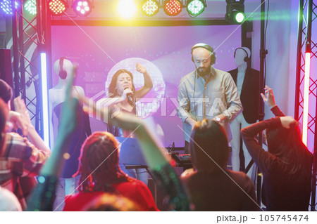 Woman singer engaging crowd to dance and performing on stage with spotlights in nightclub. People enjoying nightlife entertainment and partying on dancefloor at concert in club Woman singer engaging crowd to dance and performing on stage with spotlights in nightclub. People enjoying nightlife entertainment and partying on dancefloor at concert in club 105745474