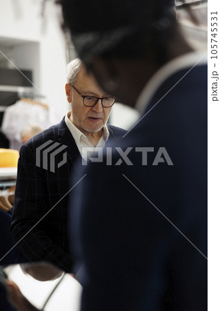 Senior client asking manager to check shirt inventory on tablet computer, wanting to buy formal wear in clothing store. Diverse people discussing merchandise fabric in modern boutique 105745531