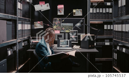 Private investigator reviewing new case evidence on detective board, conducting investigation. Woman officer examining surveillance photos and criminal records, using case files for justice. Private investigator reviewing new case evidence on detective board, conducting investigation. Woman officer examining surveillance photos and criminal records, using case files for justice. 105745537