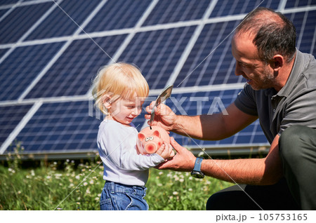 Modern father and his little son saving money in piggy bank. Adorable kid learning how to save money. Man and his cute child putting some cash to piggy bank on background of solar panels. 105753165