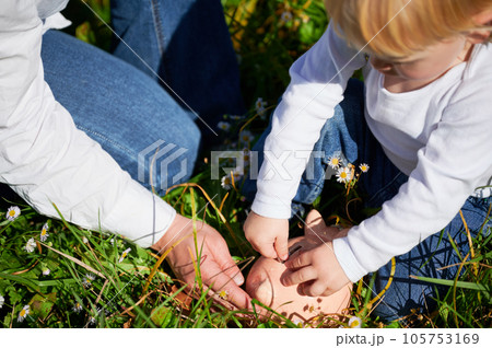 Son and young mom saving money together, putting cash into piggy bank. Mother teaching child to save money for future. Modern mom and son putting their savings into piggybank on green grass. Son and young mom saving money together, putting cash into piggy bank. Mother teaching child to save money for future. Modern mom and son putting their savings into piggybank on green grass. 105753169
