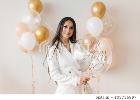 Positive woman celebrating anniversary dressed in stylish feather dress posing with birthday balloon around inflated balloons at home 105756997