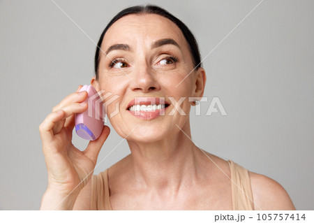 Face hand massage. Attractive mature woman looking away using silicone cleansing facial brush isolated grey background. 105757414