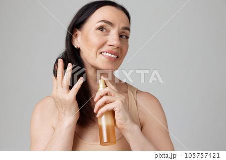 Portrait of beautiful mature brunette woman sprays remedy on hair over grey background. Hair care routine. Silky hair. 105757421