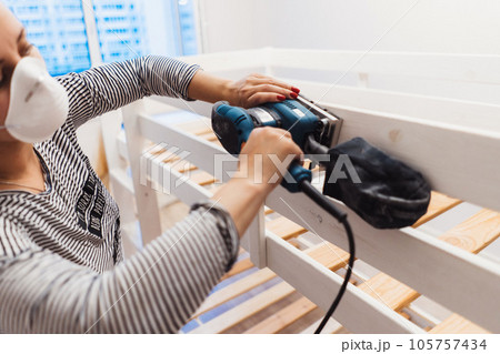 woman rubs wooden child bed with power sander or grinder removing old paint 105757434