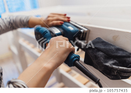 close up of woman hands rubbing wooden child bed with power sander or grinder removing old paint. 105757435