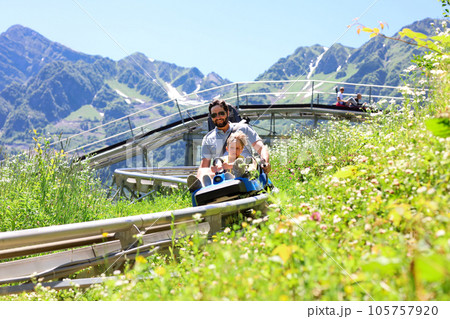 father and child having ride on summer toboggan called Rodelbahn rushing down the track . Beautiful mountains on background father and child having ride on summer toboggan called Rodelbahn rushing down the track . Beautiful mountains on background 105757920