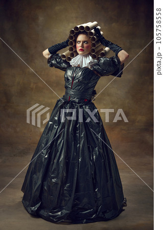 Full body portrait of elegance woman straightens hair out of paper tubs posing in dress made of garbage bags against brown background. Full body portrait of elegance woman straightens hair out of paper tubs posing in dress made of garbage bags against brown background. 105758558