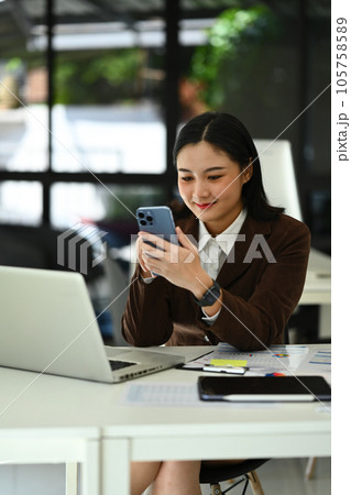 Charming asian woman entrepreneur sitting in bright home office and using smartphone. Business, technology and people 105758589