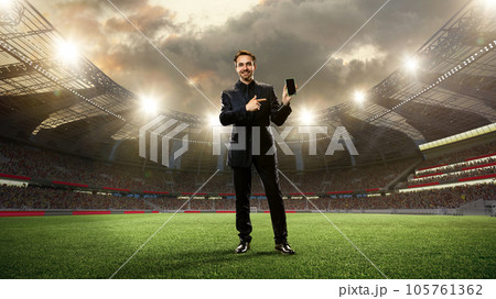 Business in a suit standing on 3d sports arena with flashlights and pointing on mobile phone screen. Betting before game 105761362