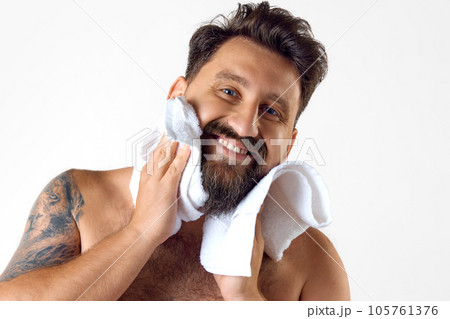 Portrait of handsome, smiling, bearded shirtless man with towel on shoulder washing face against white studio background 105761376