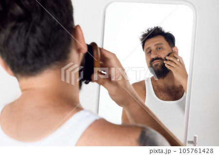 Mature man looking in mirror, taking care after beard, brushing with special brush against white studio background. Mature man looking in mirror, taking care after beard, brushing with special brush against white studio background. 105761428