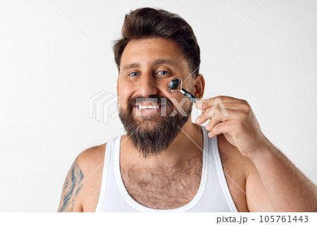 Smiling, bearded man doing face massage with roller against white studio background. Removing puffiness 105761443