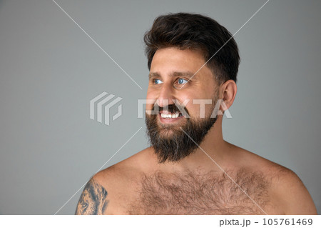 Handsome, smiling, mature, bearded man with blue eyes standing shirtless, looking away against grey studio background 105761469