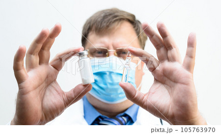 Male doctor in a mask holds an ampoule of a medical drug, close-up Male doctor in a mask holds an ampoule of a medical drug, close-up 105763970
