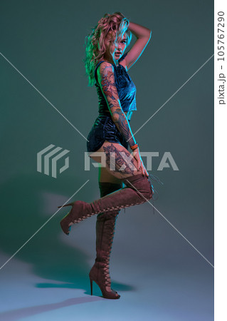 Attractive young blonde girl in blue velour booty shorts and maroon suede high boots posing on camera in studio 105767290