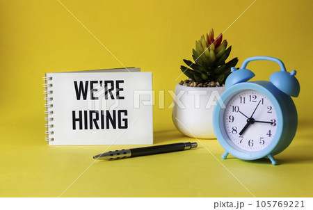 Notepad on a yellow background with the text We are hiring, next to a pen, a clock and a cactus 105769221