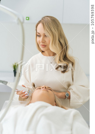 Vertical shot of female doctor performing non-invasive RF facial lifting procedure on woman for rejuvenation and skin tightening. Concept of technology hardware for anti age-related changes of face Vertical shot of female doctor performing non-invasive RF facial lifting procedure on woman for rejuvenation and skin tightening. Concept of technology hardware for anti age-related changes of face 105769398