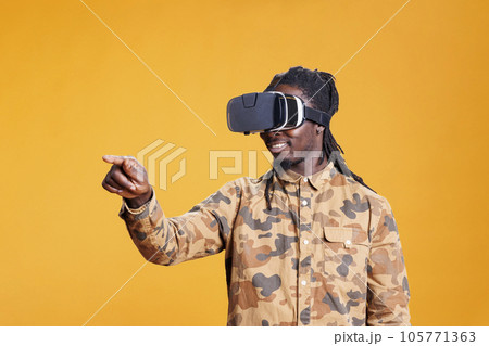 African american man using vr glasses for 3d simulation, enjoying virtual reality using interactive headset. Cheerful young adult having fun with goggles in studio over yellow background African american man using vr glasses for 3d simulation, enjoying virtual reality using interactive headset. Cheerful young adult having fun with goggles in studio over yellow background 105771363