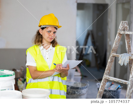 Woman foreman in a protective helmet and yellow vest checks the completed construction work on drawing 105774337