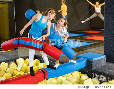 Smiling positive girls in sportswear having funny wrestling by inflatable logs in indoor game trampoline club 105774424