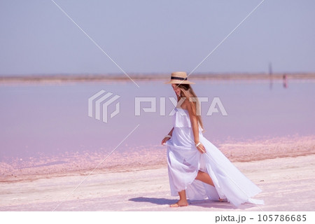Woman in pink salt lake. She in a white dress and hat enjoys the scenic view of a pink salt lake as she walks along the white, salty shore, creating a lasting memory. 105786685
