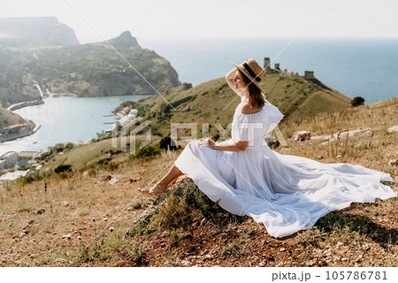 Happy woman in a white dress and hat stands on a rocky cliff above the sea, with the beautiful silhouette of hills in thick fog in the background. Happy woman in a white dress and hat stands on a rocky cliff above the sea, with the beautiful silhouette of hills in thick fog in the background. 105786781