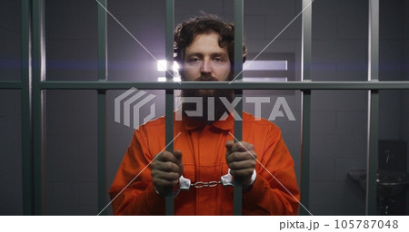 Prisoner stands in prison cell in handcuffs, looks at camera Prisoner stands in prison cell in handcuffs, looks at camera 105787048