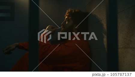 Depressed male prisoner sits on the bed, smokes cigarette in prison cell 105787137