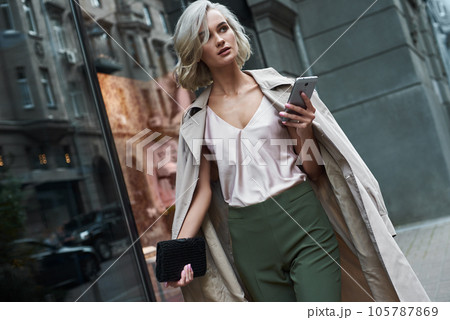 Fashion. Young stylish woman walking on the city street browsing internet on smartphone looking aside pensive 105787869