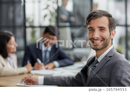 young man posing for the camera while sitting at a table in the office against the background of his colleagues young man posing for the camera while sitting at a table in the office against the background of his colleagues 105794553