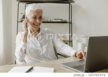 Smiling stylish mature woman sitting at desk with laptop and talking on mobile phone. 105795474