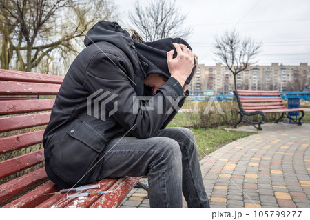 Drug abuse concept. Addict in the hood sitting on a bench in the park. Life style Drug abuse concept. Addict in the hood sitting on a bench in the park. Life style 105799277