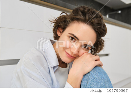 Close up portrait of cute, feminine young woman, sitting on floor, smiling and gazing at camera 105799367