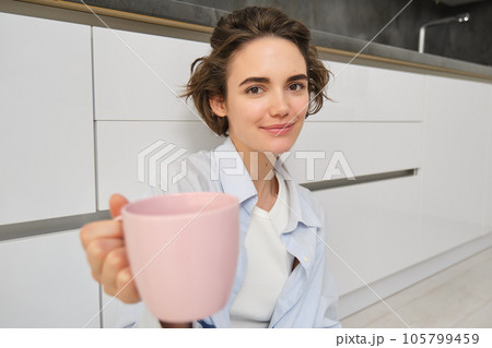 Portrait of beautiful brunette girl, gives you mug with coffee or tea, offers to drink matcha with her, holds pink cup, sits on kitchen floor and smiles at camera 105799459