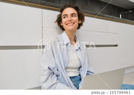 Freelance and lifestyle concept. Young woman sits on floor with laptop, works from home, sets up her remote workspace in kitchen, does homework Freelance and lifestyle concept. Young woman sits on floor with laptop, works from home, sets up her remote workspace in kitchen, does homework 105799461