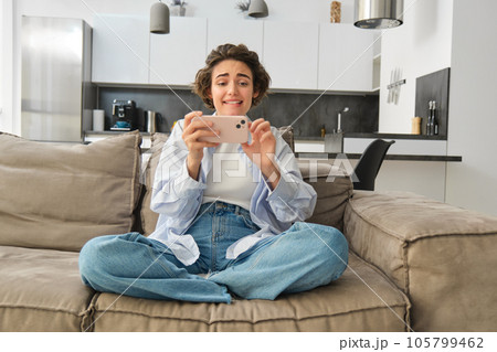 Portrait of woman playing mobile phone games, watching video with excited face, using smartphone for leisure, sits on sofa at home Portrait of woman playing mobile phone games, watching video with excited face, using smartphone for leisure, sits on sofa at home 105799462