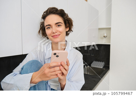 Portrait of smiling beautiful woman, sitting in kitchen with smartphone, looking happy at camera, spending time at home, using mobile phone app Portrait of smiling beautiful woman, sitting in kitchen with smartphone, looking happy at camera, spending time at home, using mobile phone app 105799467