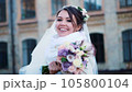 portrait of a beautiful bride, in a white dress, veil and fur coat. bride is smiling happily, holding a wedding bouquet. against the background of ancient architecture, winter wedding. 105800104