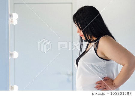 Black-haired chubby woman in t-shirt overweight lady looks at reflection on mirror at home 105801597