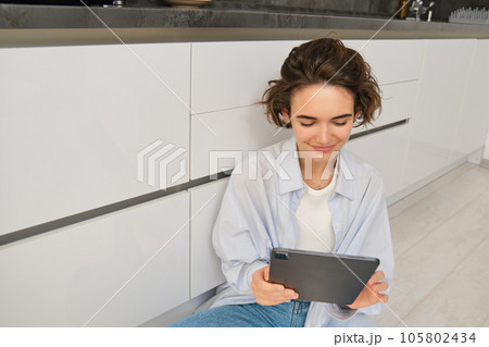 Portrait of woman looking at her tablet, reading online, watching videos on digital device, sitting on floor at home Portrait of woman looking at her tablet, reading online, watching videos on digital device, sitting on floor at home 105802434
