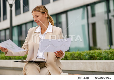 Stylish woman analyst working with documents while sitting outside on modern building background Stylish woman analyst working with documents while sitting outside on modern building background 105805493