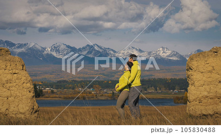 Man and woman in yellow green sportswear. Lovely couple of travelers hug and kiss near old stone enjoying highland landscape. 105830488