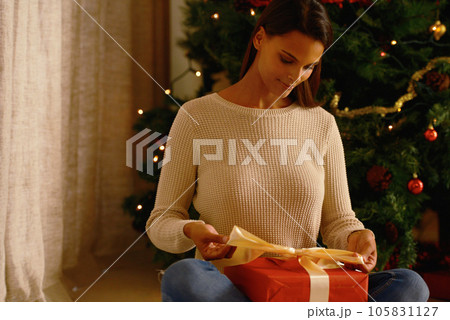 Christmas, ribbon and woman with a gift by a tree wrapping a box or present on a Christian holiday at home. Night, relax and happy person enjoys giving presents in bow decoration in a house in winter Christmas, ribbon and woman with a gift by a tree wrapping a box or present on a Christian holiday at home. Night, relax and happy person enjoys giving presents in bow decoration in a house in winter 105831127